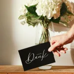 Schlichte Schwarz-Weiß Dankeskarte in einer Hand vor Blumenstrauß - A6 Karte mit 'Danke für deine Bestellung' und Kontaktdaten