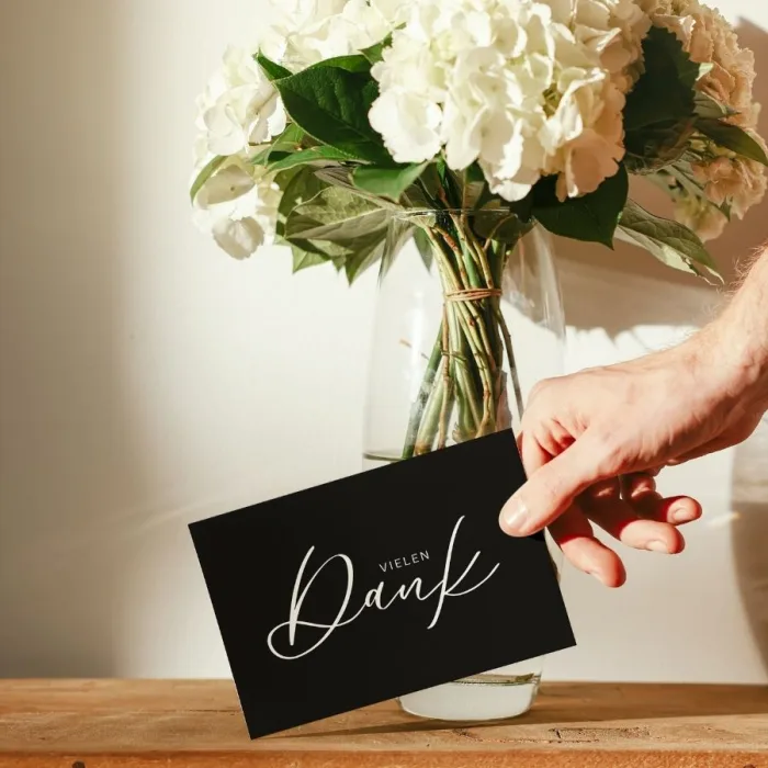 Schlichte Schwarz-Weiß Dankeskarte in einer Hand vor Blumenstrauß - A6 Karte mit 'Danke für deine Bestellung' und Kontaktdaten