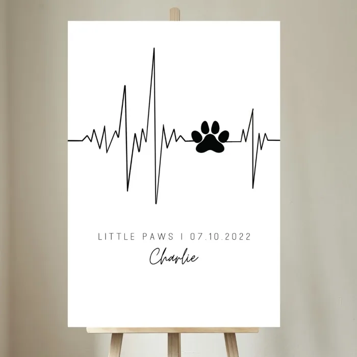 Dieses Bild zeigt das personalisierte 'Little Paws' Bild mit dem Namen und Geburtsdatum des Hundes sowie einer Herzschlagzeichnung in einem gemütlichen Wohnraum. Das moderne Design fügt sich perfekt in die warme und einladende Atmosphäre ein und ist eine ideale Wanddekoration für Hundeliebhaber, die ihre Liebe zu ihrem Vierbeiner ausdrücken möchten.