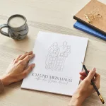 Unerahmtes Familien-Poster im Line-Art Stil mit Bibelvers auf Schreibtisch, daneben Frau mit Bleistift